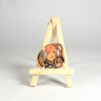 Broche brodée dans les tons orangés, gris et argent. Des volutes formées par une soutache orange. Les creux sont remplis de coquillages, perles de verre soufflé argent, et de fils. Des cupules en laines feutrée rebrodée de coton. Des perles de rocaille apporte un peu de brillance à l'ensemble.