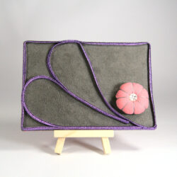 Pâté de broderie posé sur un chevalet. Le tissu de fond est gris et la soutache qui en fait tout les tour est violette. Un coeur en soutache compartimente le pâté. Un petit coussinet à aiguilles est fixé dans le bas à droite.