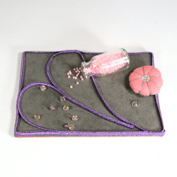 Pâté de broderie. Fond gris, soutache violette. Une fiole déverse des perles roses, assortie au coussinet.