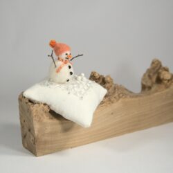 Bonhomme de neige en laine feutrée rebrodée.Son bonnet à pompon, son nez et son écharpe sont oranges. Il est fixé sur un coussin blanc dont le dessus est brodé de rubans et de perles. Le coussin est posée sur une loupe de bois