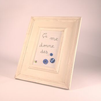 Broderie encadrée. L'expression "Ça me donne des boutons " est brodée au point de chaînette à main levée en noir sur fond blanc. Les boutons sont bleus. Cette broderie peut s'accrocher au mur ou simplement être posée sur un meuble. Vue légèrement de côté.